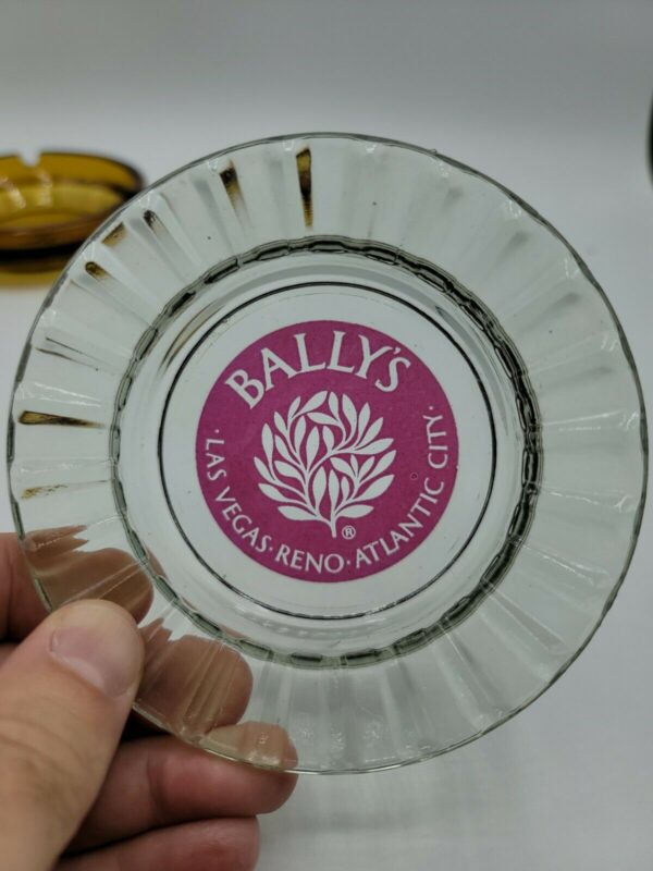 A person holding a Vintage Las Vegas Casino Ashtrays Bally's Castaways Peppermill Hilton glass plate with a logo on it.