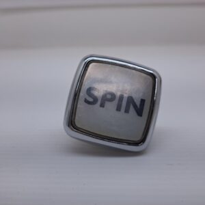 A Konami Gaming "Spin" Button with rounded edges and Chrome (plastic/coated) finish. NEW. GETT Part BTN217 with the word spin on it.