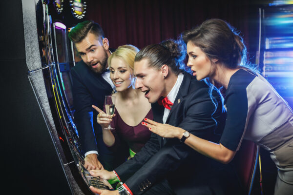 Four people excitedly playing slot machines in a casino, holding drinks and enthusiastically reacting to the FLUORO Ballast for IGT.  IGT 195 076 90. 25voc, 0.62A GETT Part Ballast105.