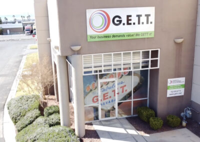 Entrance of a building with a sign "g.e.t.t. your business demands value! we gett it" above the glass door, flanked by bushes, under a clear sky.